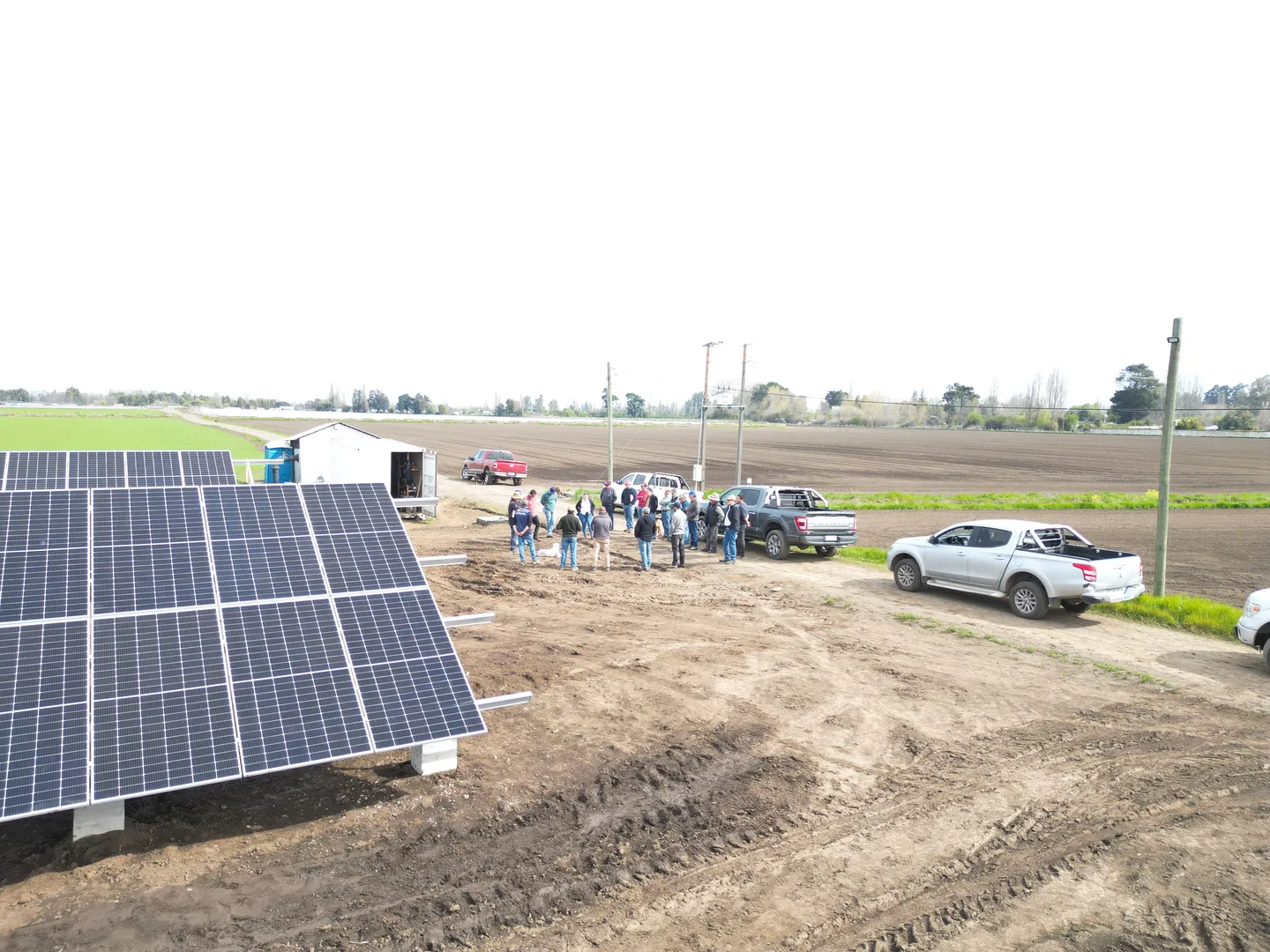 Vista aérea de una planta fotovoltaica sobre suelo junto a un contenedor técnico; un grupo de personas conversa en el camino de acceso, junto a camionetas, con campos de cultivo al fondo.