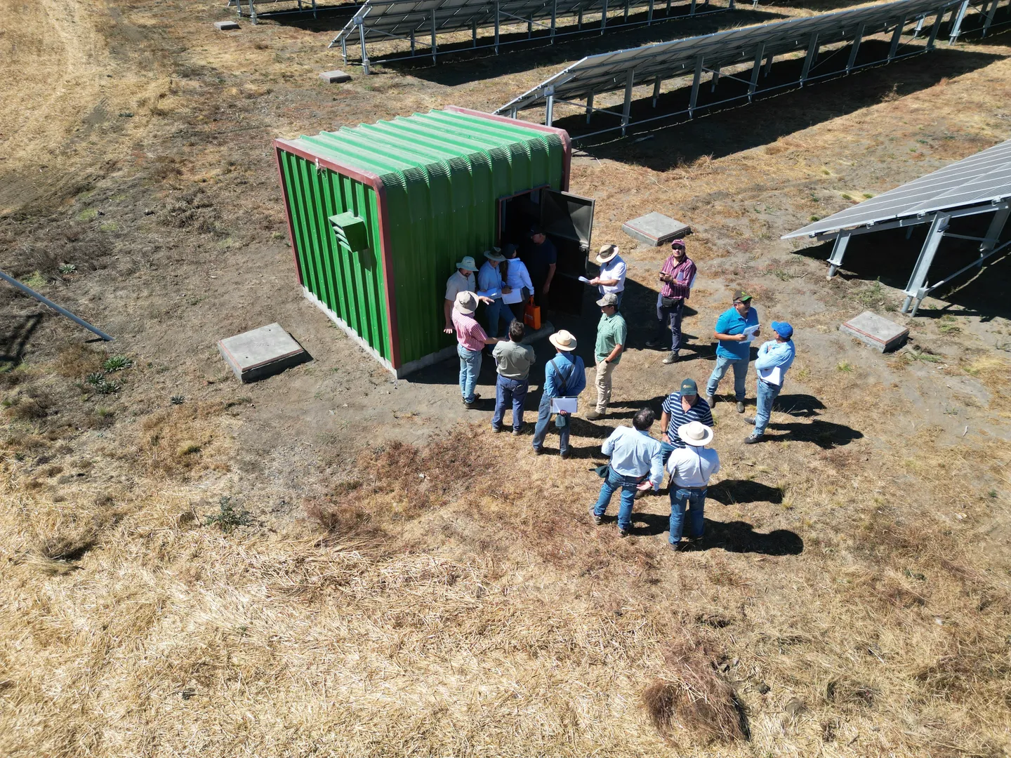 Vista aérea de un grupo de personas junto a una caseta de bombeo verde, rodeada de filas de paneles fotovoltaicos sobre suelo, en un campo de pasto seco.