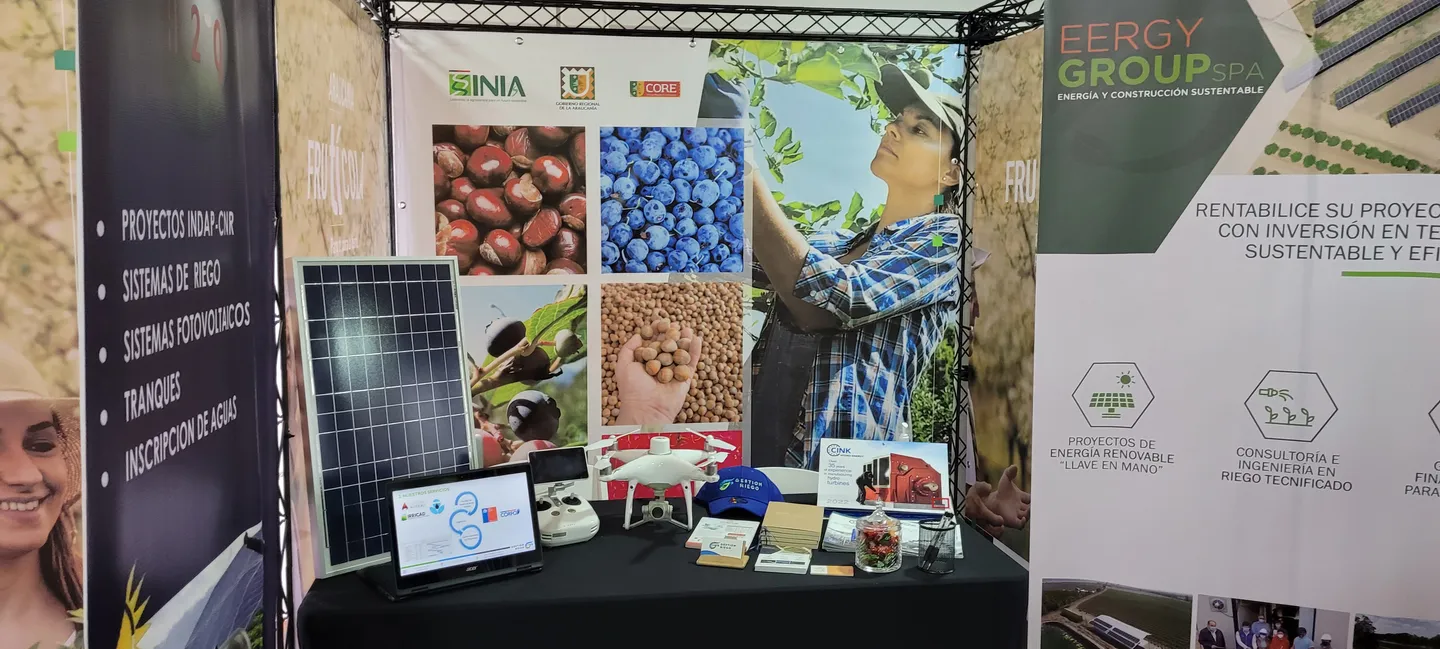 Stand con una mesa de exhibición: material impreso y una gorra de Gestión Riego, un dron, un computador portátil y un panel solar, entre gráficas de la feria y un pendón de EERGYGROUP.