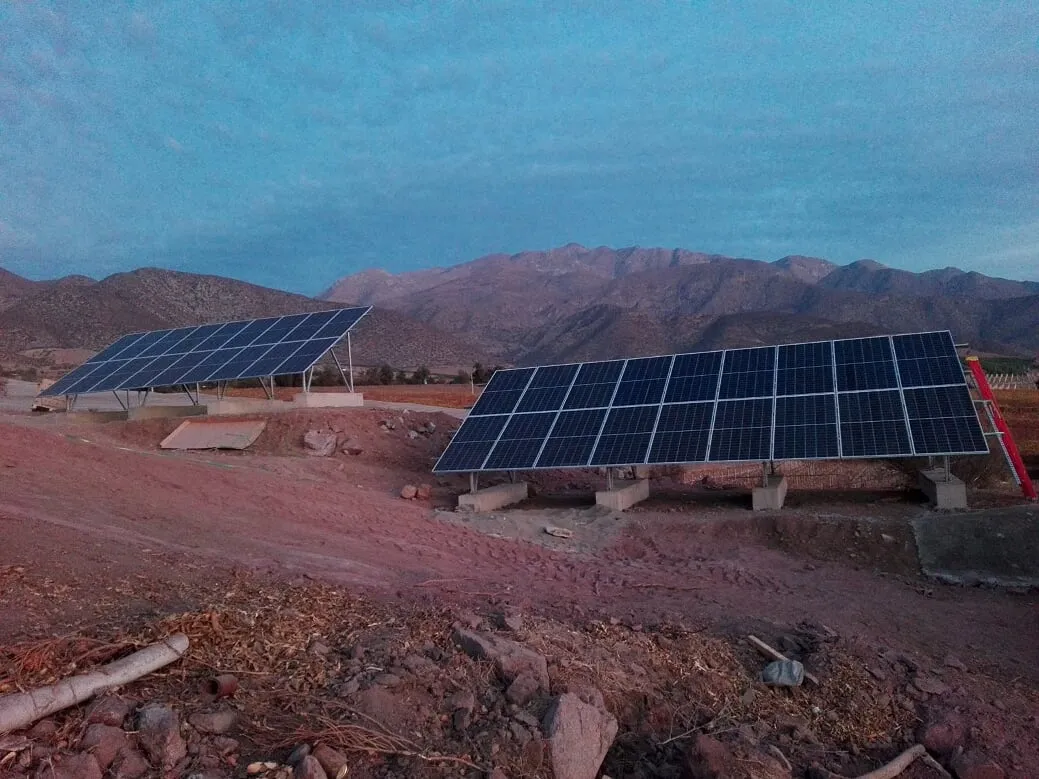 Dos mesas de paneles fotovoltaicos sobre bases de hormigón, en terreno pedregoso al pie de cerros áridos.