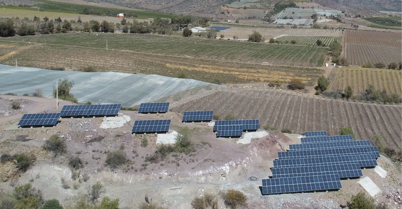 Mesas de paneles fotovoltaicos sobre plataformas cortadas en una ladera seca, por encima de cultivos en hileras y un sector con malla.