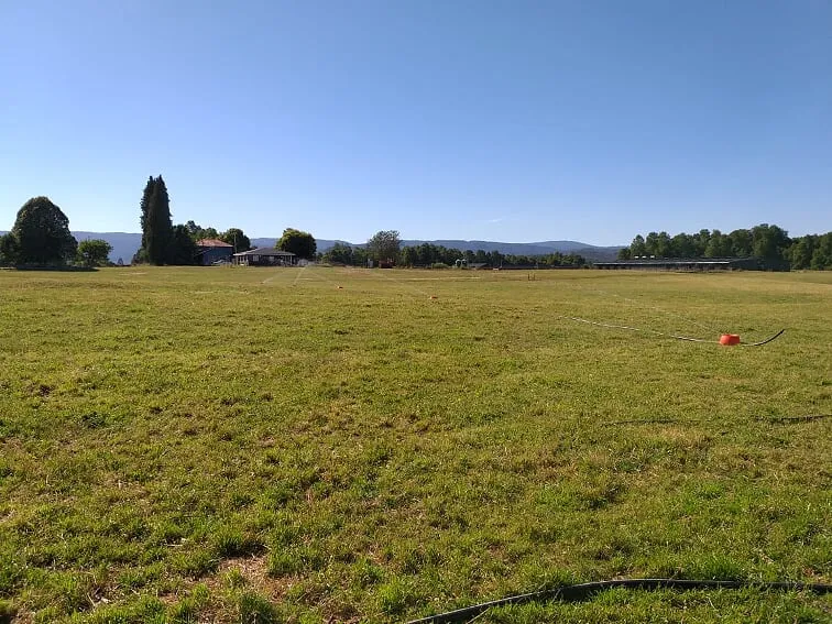 Pradera plana con tazones naranjos unidos por manguera negra; al fondo, casas, un galpón largo y cerros con bosque.