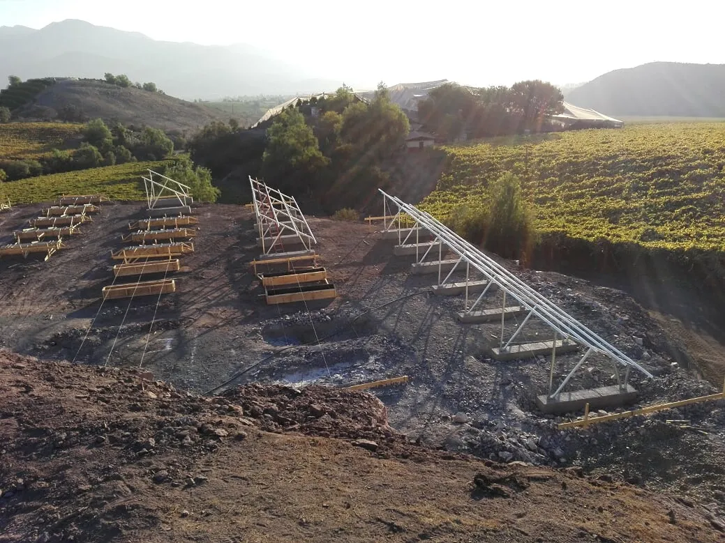 Estructuras metálicas triangulares sobre bases de hormigón, moldajes de madera y lienzas de trazado junto a cultivos en hileras.