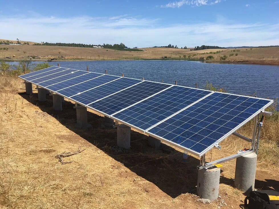 Fila de paneles fotovoltaicos sobre estructura de aluminio y pilares de hormigón, a orillas de un tranque entre lomajes secos.