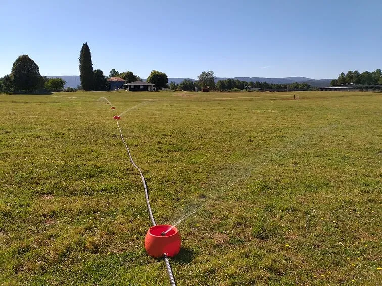 Aspersor de tazón naranjo lanzando agua sobre una pradera plana; la manguera sigue hacia otros tazones de la línea.